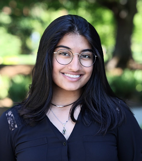 South Asian woman with glasses and long dark hair smiles at the camera; greenery and a tree blurred in backtround