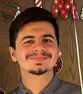 Young white man with short dark hair, a moustache and goatee, wearing a grey collared shirt smiles in front of a wooden surface with the letters S, I and M visible