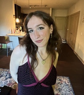 Young white woman with long brown hair wearing a sundress and necklace smiles at the camera; a stool, carpet, light, and closet visible in the background