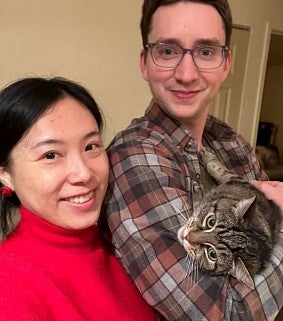 Daisy in a red sweater next to her husband in a checked shirt, holding their tabby cat, Edamame