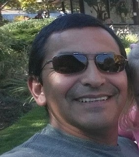 Man with short black hair and sunglasses wearing a grey T-shirt; behind him are sunlight, grass, bushes, and trees.