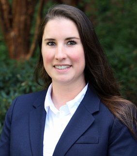 White woman with long brown hair, wearing a white collared shirt and blue blazer smiles at the camera