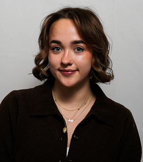 Young white woman with brown shoulder-length hair in a brown collared shirt smiles into the camera; light grey background