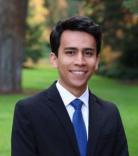 Young Hispanic man in a suit and tie smiles confidently; grass and trees in the background