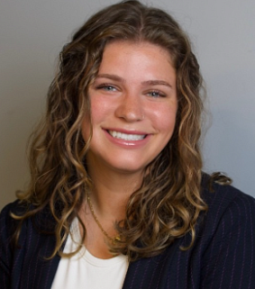 Young white woman with brown hair past her shoulders and a blue blazer smiles; grey wall behind her