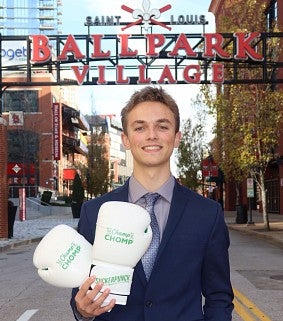 Young white male in a suit and tie holds boxing gloves imprinted with "The Champ of Chomp"; behind him lettering reads Saint Louis Ballpark Village