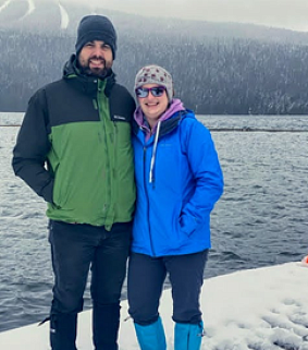 White man and woman wearing woolly hats and winter clothing stand on snow next to a dark lake; trees in background