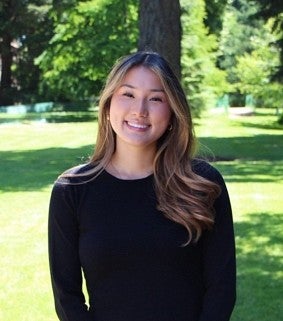 Young Asian woman with long light brown hair and a black shirt smiles at the camera; trees and grass in the background