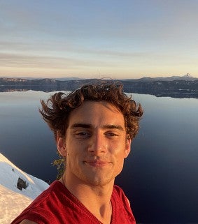 Young white man with wavy brown hair and a red top on looks at the camera; the sun is shining on his face from right; in the background is snow and a lake