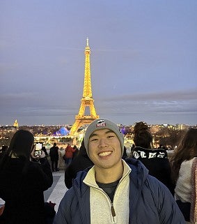 Young Asian man in warm clothes and beanie smiling in front of a night sky; in the background is the Eiffel Tower, lit up in orange/yellow