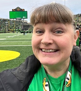 White woman with brown hair tied back; standing on the field at Autzen stadium; wearing a green UO Ducks shirt and black jacket