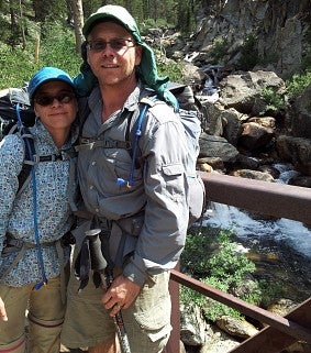 Woman and man--both wearing hiking gear and backpacks pose on a bridge in front of a waterfall