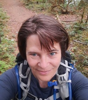 White woman with short brown hair wearing a blue shirt and hydration backpack; trail and trees in background