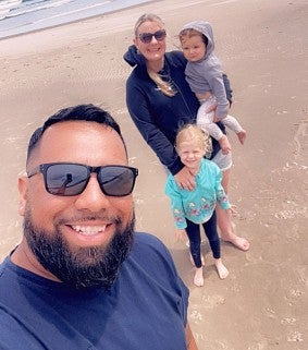 Eric, his wife, and two daughters at the beach--standing on the sand with the ocean behind them