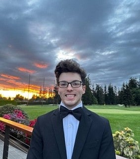 Young white man with dark hair and glasses wearing a bow tie and tuxedo; behind him are lush gardens, a cloudy sky, and sunset