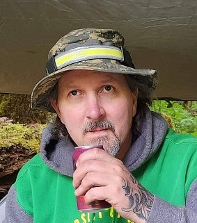 White man with moustache and goatie wearing a camouflage hat and green-grey hoodie raises a soda can to his mouth; he is outdoors; greenery in the background and a tarp above his head