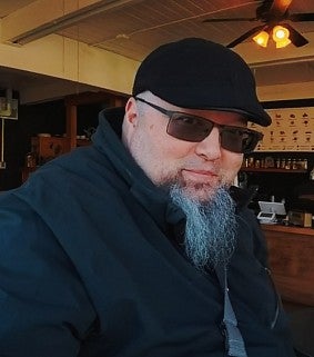 Asian-American man with a stylish grey beard in a black ball cap and sunglasses looks into the camera; coffee shop in the background