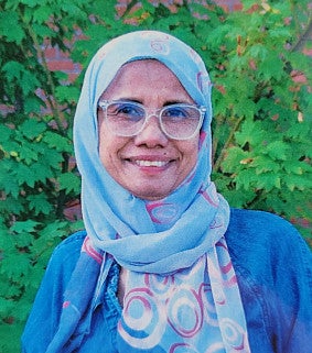 Woman wearing glasses, a headscarf, and blue top smiles at the camera; a red brick building and green foliage are visible behind her 