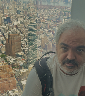 Man with short grey hair, moustache, and beard looks at the camera; behind him is the Jenga Tower in New York
