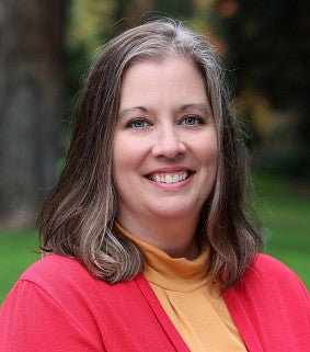 A white woman with shoulder-length brown hair smiles at the camera; there is a tree, green grass and foliage behind her