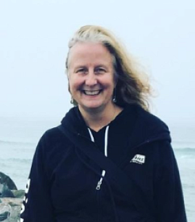 White woman with long blonde hair smiles in front of the ocean