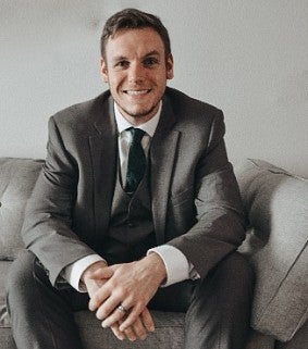 Man in a grey suit sits on a couch with his hands together, smiling at the camera