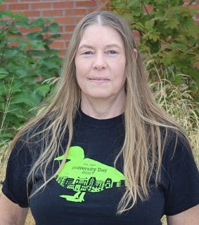 Woman with long sandy blonde hair in a black t-shirt. Red brick and green leaves.
