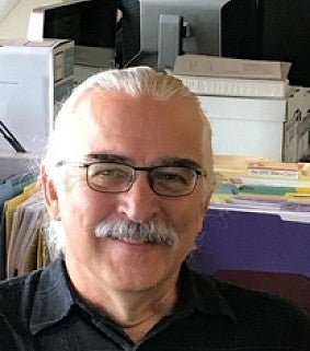 A man with glasses and a mustache sits in an office environment with folders, boxes, and computer screens behind him