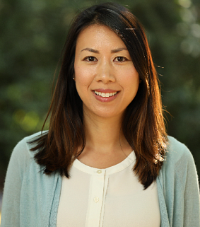 A woman with long hair smiles at the camera; green foliage visible in the background