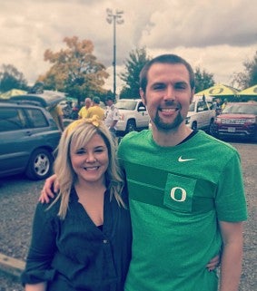 A woman with a yellow ribbon in her long, blonde hair smiles on the left of the photo; on the right, next to her is a man wearing a UO shirt, also smiling