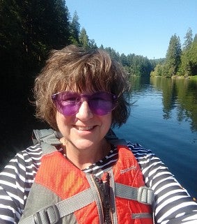 Woman with short brown hair, sunglasses, long-sleeved shirt and life jacket smiles; background is calm water, blue skies, and green trees