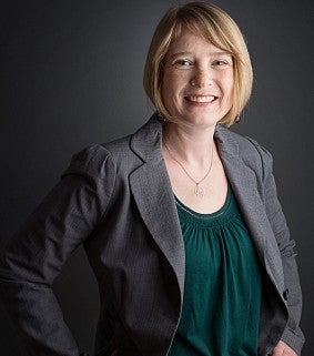 A woman with blonde hair framing her face smiles at the camera; she is wearing a green top and grey jacket