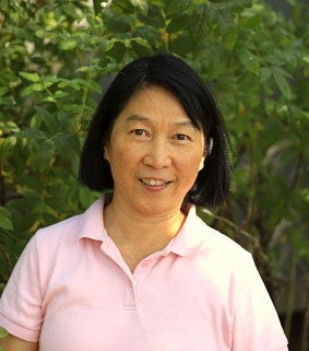 A woman with dark hair and a pink shirt smiles at the camera; green foliage if visible in the background