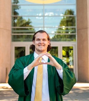 Man with shoulder-length hair and a green graduation gown makes the "O" sign