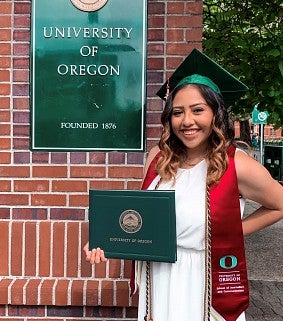 Woman with long, brown hair in a white dress and green graduation gown in front of a UO sign