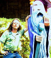 Man with long brown hair, floral shirt, jeans, and hand on his hip leans on the shoulder of a statue--of a sage with a long grey beard and staff. Flowers visible behind the man.