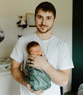 Young white man with short dark hair, moustache, and short beard holds a small baby wrapped in green