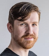 White man with short brown hair, ginger moustache and short beard and wearing a black T-shirt looks at the camera; white background