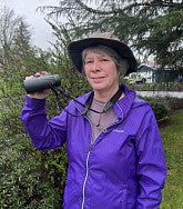 Woman with pale complexion, short grey hair, brown hat and purple jacket; holding a pair of binoculars; grass, trees, and a bush behind her