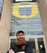 White man with brown hair, beard, and moustache, black cap and "O" Facilities Services vest smiles broadly into the camera; arms folded; class doors and windows and big yellow O behind him
