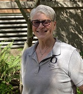 White woman with short grey hair, black rimmed glasses, wearing a light grey collared O Facilities Services, short-sleeved shirt smiles into the camera; behind her is a tree with leaves casting dappled shadows on an exterior wall