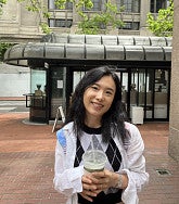 Asian woman with black hair past her shoulders smiles at the camera; she is holding a drink; in the background is a coffee shop converted from an old bus stop