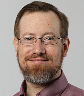 White man with short light brown hair, beard with hints of grey, a moustache and glasses; wearing a light burgundy collared shirt; white background; smiles into camera