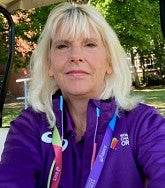 White woman with shoulder length blonde hair looks into the camera; she is wearing a purple OR22 top; behind her are green grass and green trees