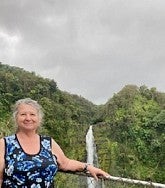 Light-skinned woman with gray hair and a sleeveless blue floral top stands with her arm on a wire fence; behind her are lush green hills and a cascading waterfall; gray skies overhead.