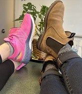 Two sets of legs and feet crossed and up on table; one set in pink sneakers, the other set in brown boots; a potted plant in the background