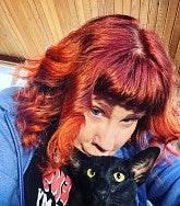 White woman with red hair looks down at the camera; she is leaning her chin on a black cat