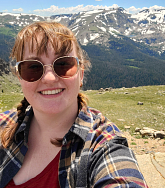 Mary with hair up, sunglasses on in front of a snow-capped mountain range; grass, rocks, blue skies and clouds also visible in the background