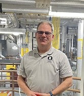 White man with glasses and a grey shirt stands in front of pipes (inside the central power station)