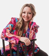 Young white woman with long light brown hair sits astride a chair facing the camera; she is wearing a pink, blue, red, and purple jacket and black trousers